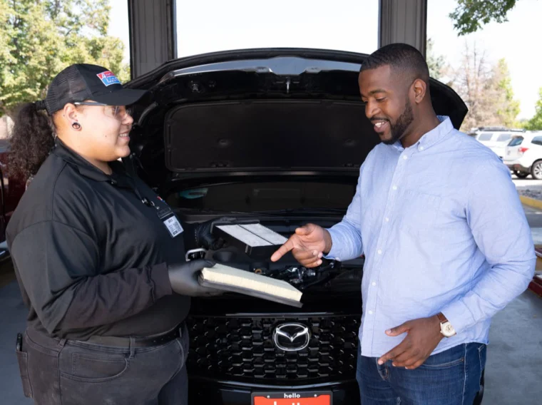 An auto technician shows an air filter to a customer in front of a car with its hood open at a service center, explaining the importance of regular air filter replacement.