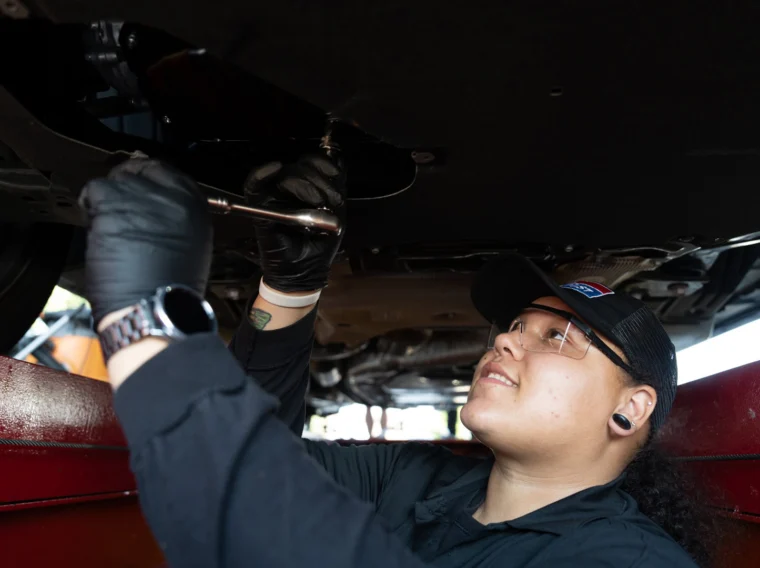 A mechanic wearing gloves and safety glasses works underneath a raised vehicle using a wrench.