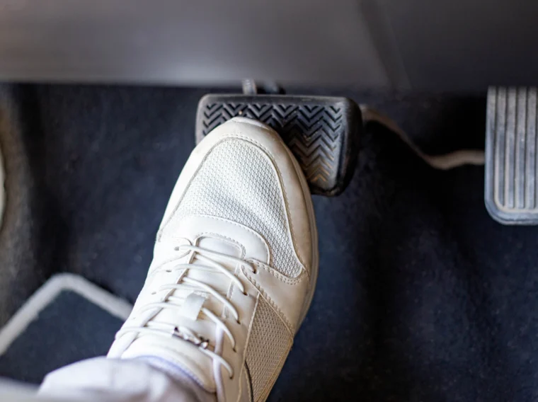 A person wearing a white sneaker presses down on a car brake pedal, with the accelerator pedal visible to the side—an essential step when performing tasks like clutch replacement or cylinder replacement.