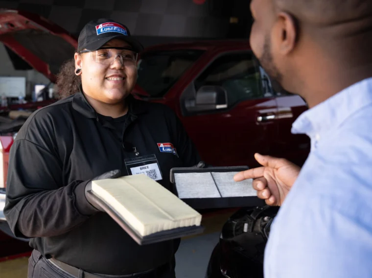 An auto technician discusses Air Filter Services with a customer, showing a comparison between a clean and dirty air filter inside a garage with vehicles in the background.
