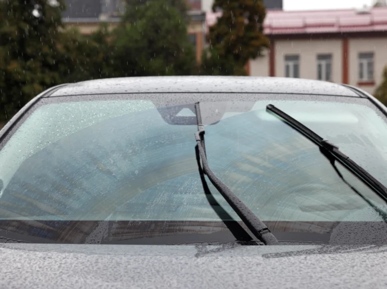 A close-up of a car windshield with two windshield wiper blades in motion, clearing rainwater while parked outdoors on a rainy day.