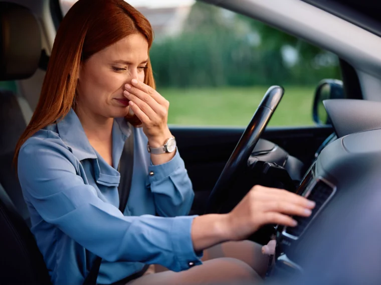 A woman sitting in a car pinches her nose with one hand and adjusts the controls with the other, seemingly reacting to an unpleasant smell—perhaps a sign that a clutch replacement or cylinder replacement is needed.