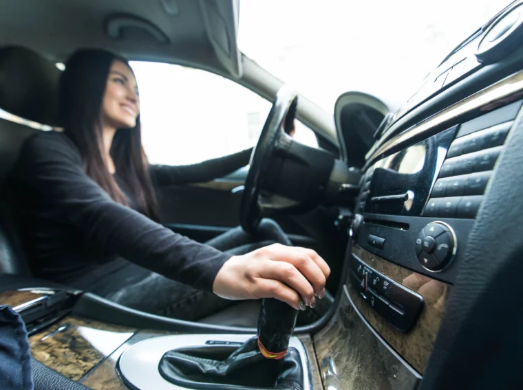 A person in the driver's seat of a car holds the steering wheel with one hand and shifts the gear lever with the other, possibly after a recent clutch replacement to ensure smooth and efficient gear changes.