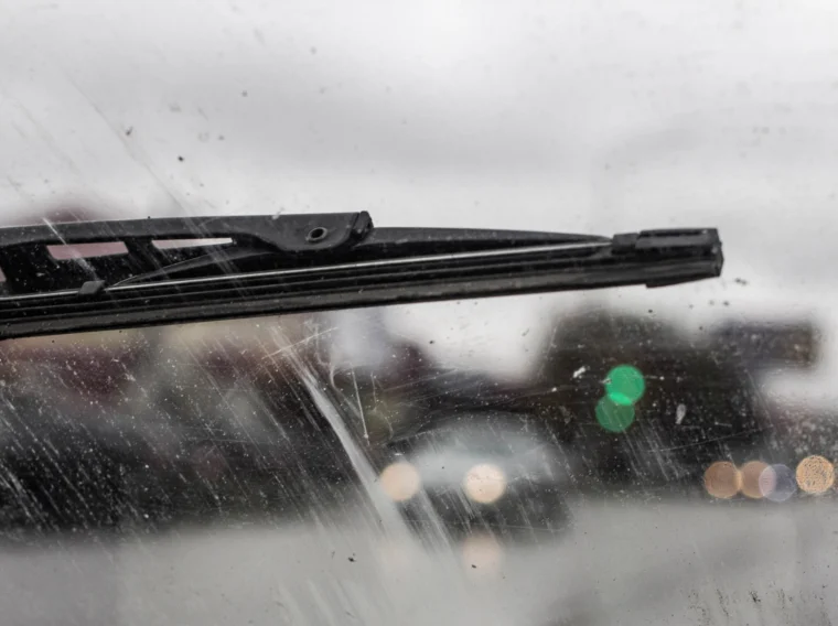 Close-up of a dirty windshield with a windshield wiper blade, blurred view of cars and traffic lights visible through the glass on a cloudy day.