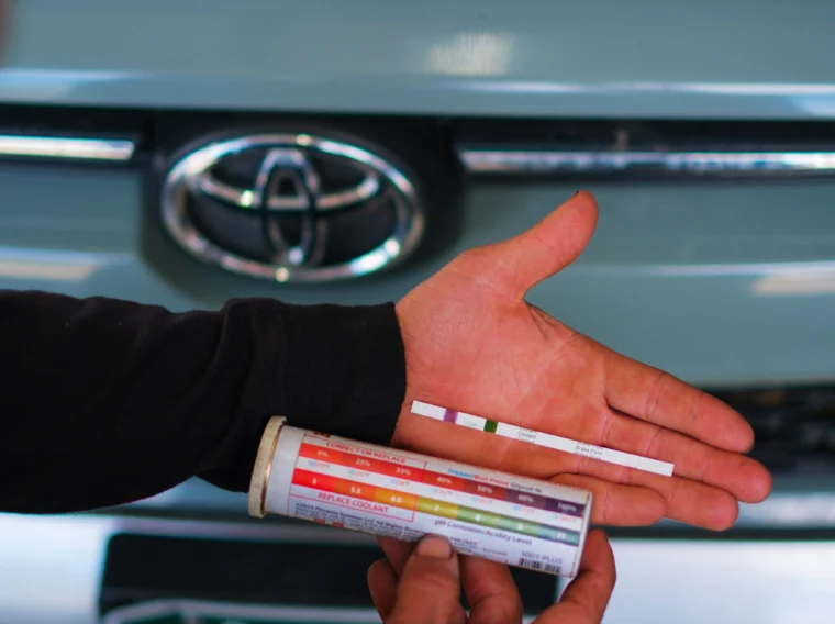 Person holding test strips and a color chart in front of a Toyota car, possibly testing automotive fluids under the hood as part of a cooling system repair.