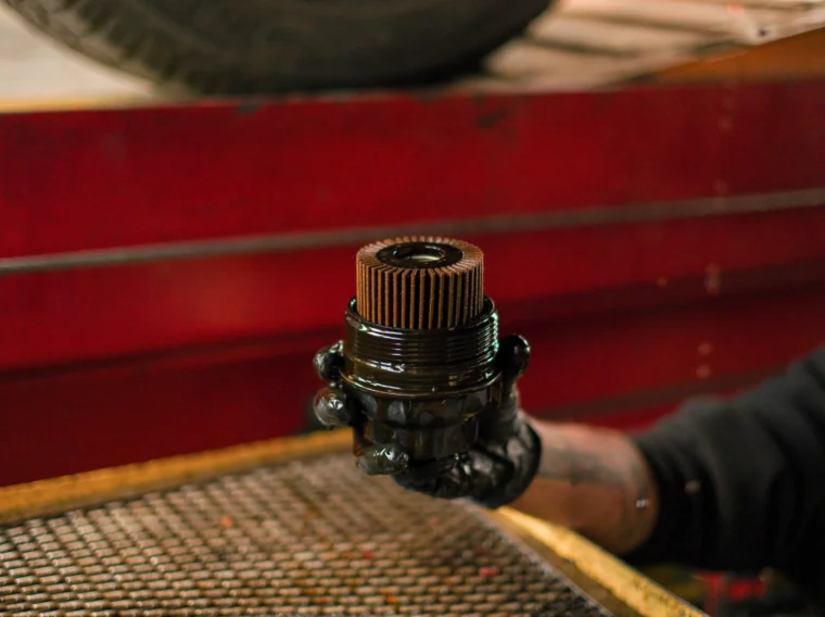 A person wearing a black glove holds a used, oil-covered engine oil filter in a garage setting, highlighting the importance of regular Filter Services.