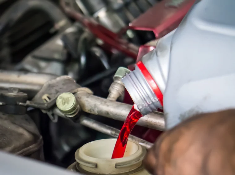 A person pours red engine fluid from a plastic bottle into a reservoir in a car engine bay.