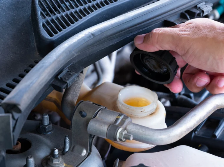 A hand holds the cap above an open brake fluid reservoir in a car engine bay, showing the fluid level inside the container.