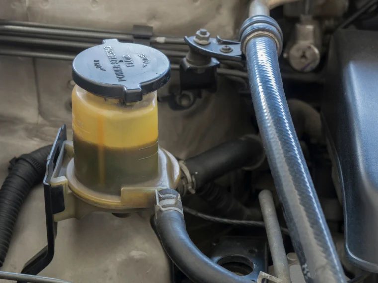 Close-up of a car engine compartment showing a brake fluid reservoir with discolored fluid, indicating possible contamination. A Power Steering Flush may also be needed to maintain overall steering system performance.