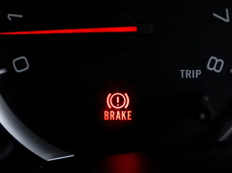 Close-up of a car dashboard showing an illuminated red brake warning light and part of the speedometer.