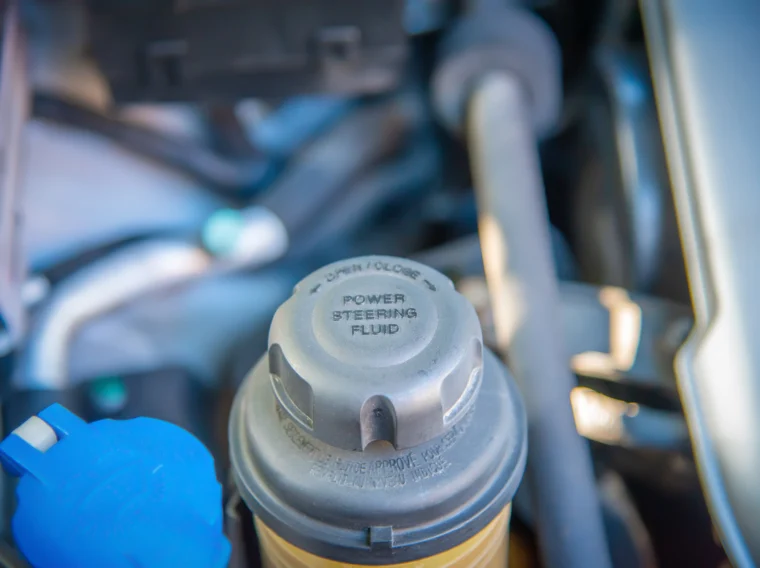 Close-up of a car engine compartment showing the cap for the power steering fluid reservoir, labeled "Power Steering Fluid," with another blue cap nearby—ideal for illustrating Power Steering Services or a Power Steering Flush.