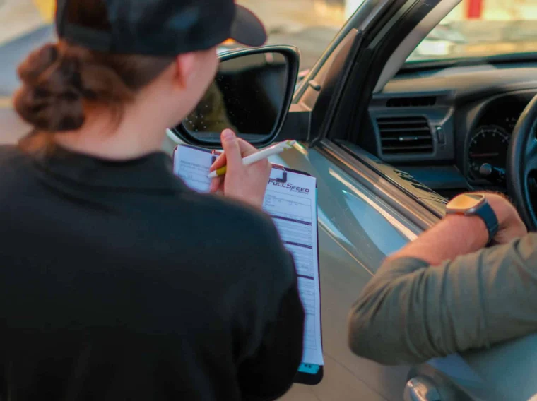 A worker holding a clipboard speaks with a person in a car at a service station, while other employees handle car cleaning tasks in the background near caution tape.