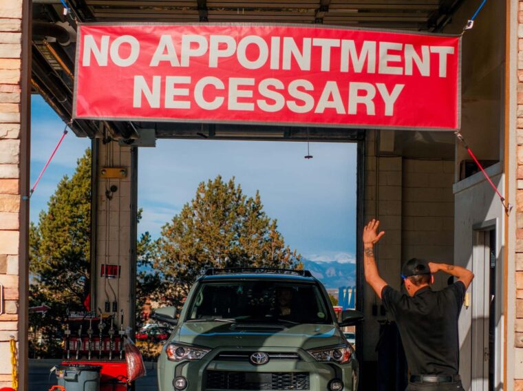 A car enters a service station under a red "No Appointment Necessary" sign while a worker in black raises his arm to guide the vehicle, ready to assist with Fuel Injection Cleaning.