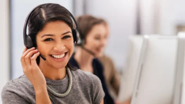 Woman wearing a headset smiles at the camera while sitting at a desk; another person with a headset works at a computer in the background.