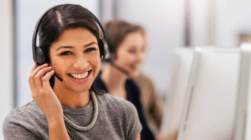 Woman wearing a headset smiles at the camera while sitting at a desk; another person with a headset works at a computer in the background.