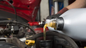 A person pours motor oil from a plastic container into a car engine with the hood open, wearing a black glove.