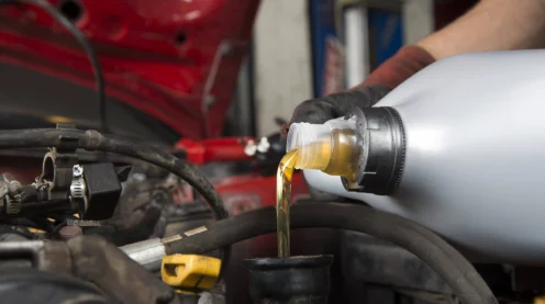 A person pours motor oil from a plastic container into a car engine with the hood open, wearing a black glove.