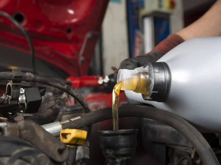 A person pours motor oil from a plastic container into a car engine with the hood open, wearing a black glove.