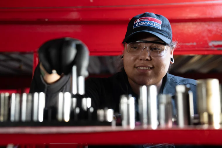 A mechanic wearing safety glasses and a "LubeFast" cap selects a tool from an organized set, with a red background.