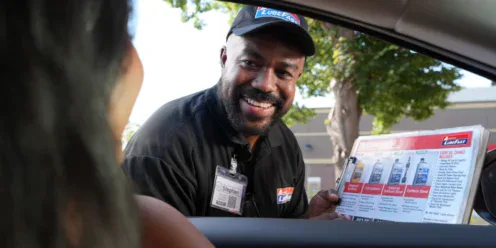 A smiling automotive employee shows an oil change options chart to a customer sitting in a car.