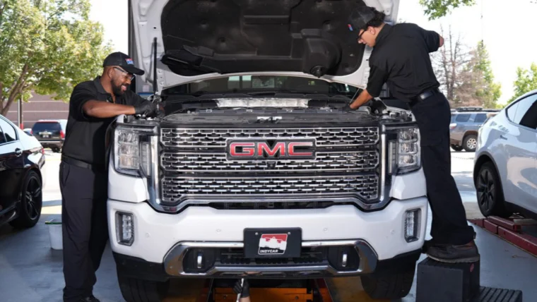Three mechanics service a white GMC pickup truck in a garage; two work under the open hood while one works beneath the vehicle in a maintenance pit.