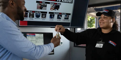 A customer receives car keys from an employee at an auto service center counter, with a screen behind them displaying tire pressure monitoring information.