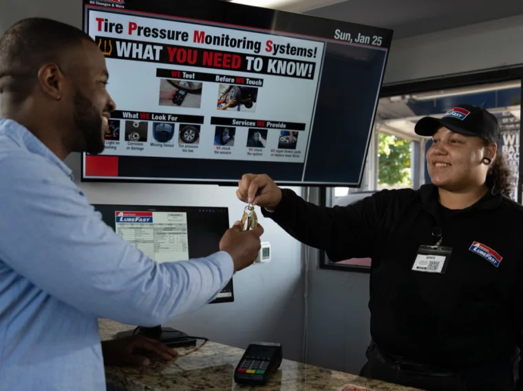 A customer receives car keys from an employee at an auto service center counter, with a screen behind them displaying tire pressure monitoring information.