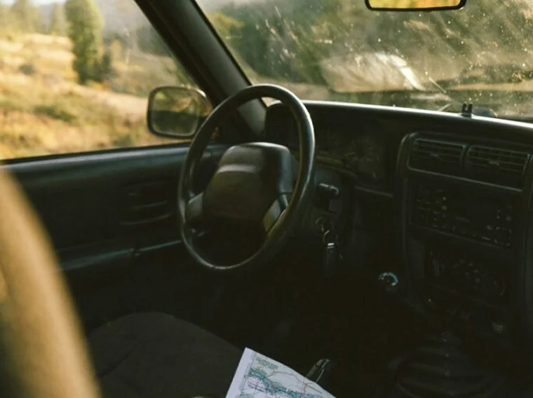 Interior of a car with the steering wheel, dashboard, and a map on the passenger seat visible; outside scenery is blurred through the windows, capturing the essence of Cars of Character on a journey.