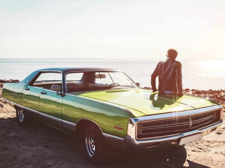 Person in sunglasses and a leather jacket leans against a green vintage car from Cars of Character, parked near a beach with water and sunlight in the background.
