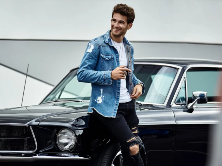 A man with character in a denim jacket and ripped black jeans leans against a vintage black car, smiling and holding sunglasses.