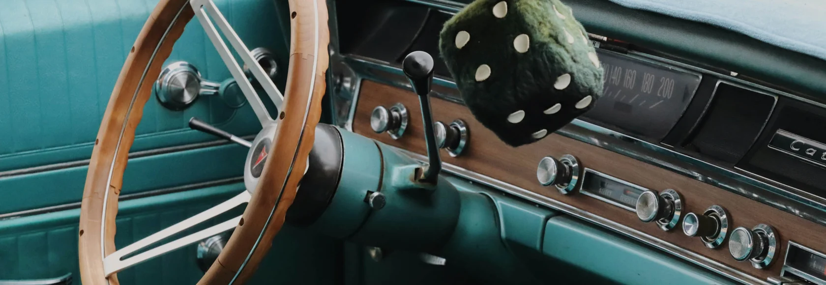 Interior of a vintage car from the Cars of Character collection, featuring a wood-rimmed steering wheel, manual gearshift, turquoise upholstery, and fuzzy dice hanging from the rearview mirror.