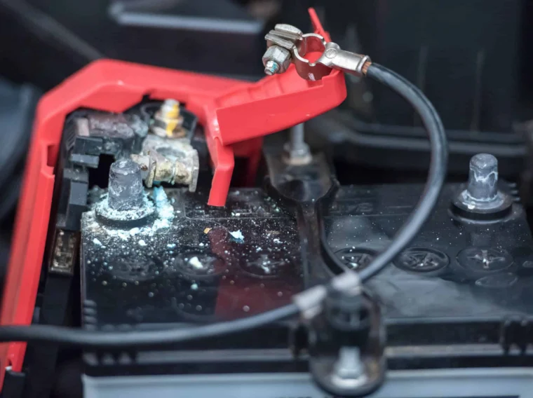 A car battery with corrosion visible on the terminal, a red protective cap is lifted, and connecting cables are attached—an example of when battery services may be needed.