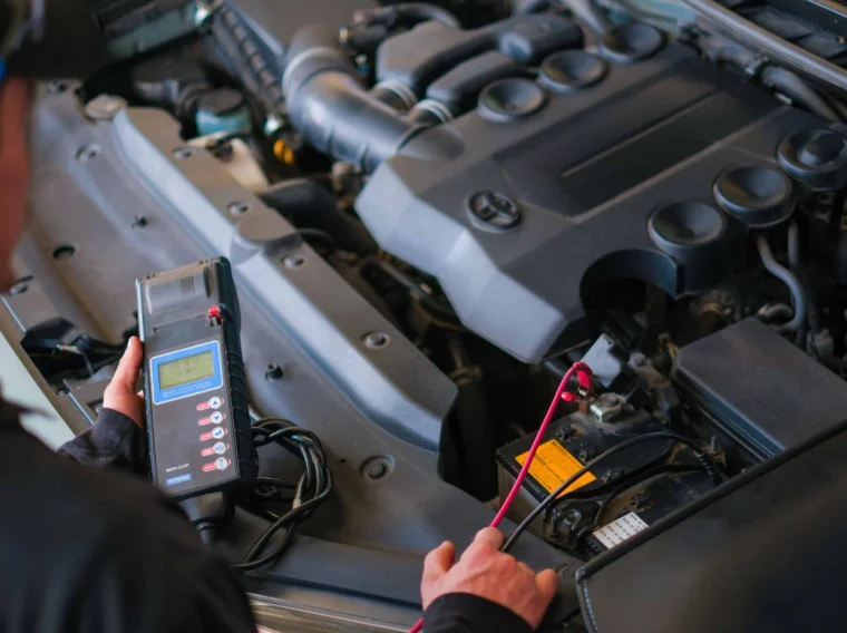 Person uses a diagnostic tool to check a car battery under the hood, with engine components visible in the background, as part of professional Battery Services.