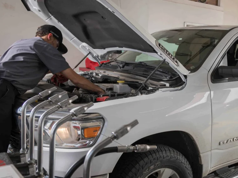 A mechanic performs battery services under the hood of a white Ram truck in a garage while a person sits in the driver’s seat.